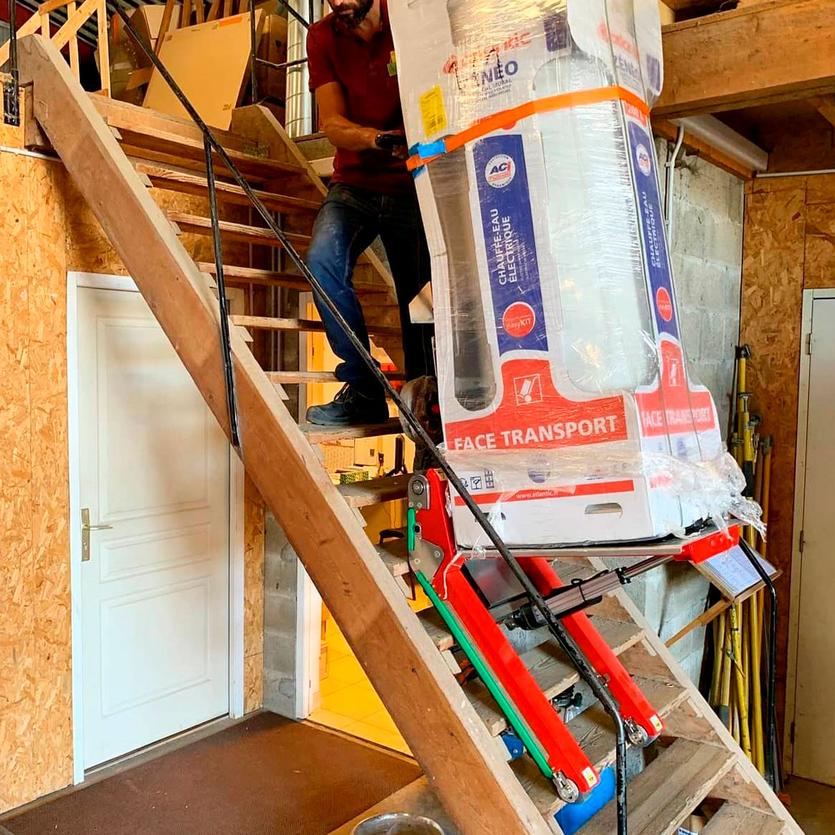 Ein Mann transportiert eine schwere Verpackung mit einem elektrischen Treppensteiger mit Raupen eine sehr steile Holztreppe hinauf
